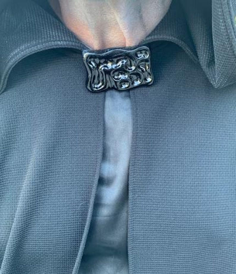 May include: A close-up of a black cloak clasp. The clasp is rectangular with an abstract, textured design. The cloak is a dark blue-grey color, with a textured weave. The background is a lighter blue-grey.