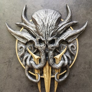 Può includere: Una scultura da parete in 3D stampata in argento e oro che rappresenta una creatura mostruosa con tentacoli e un viso simile a un teschio. La creatura è circondata da una cornice circolare con un bordo dorato.