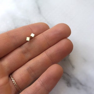 May include: A pair of small, gold square stud earrings. The earrings are shown on a person's hand.