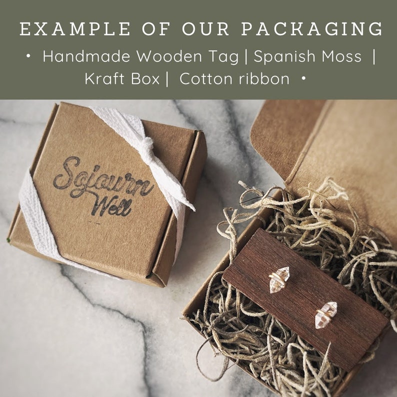 May include: A small brown box with a white ribbon tied around it. The box is labeled "Sojourn Well". The box is open and filled with Spanish moss. A small wooden tag with two clear crystal earrings is sitting on top of the moss.