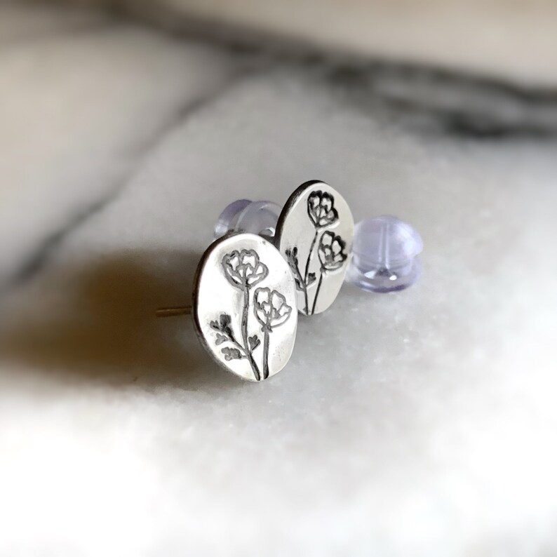 May include: A pair of silver stud earrings with a floral design. The earrings are oval shaped and feature a delicate outline of three flowers.