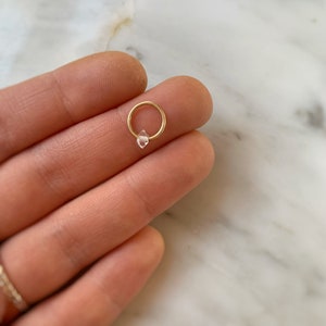 Op de afbeelding: Een gouden oorring met een heldere kristallen edelsteen. De oorring wordt getoond op een hand.