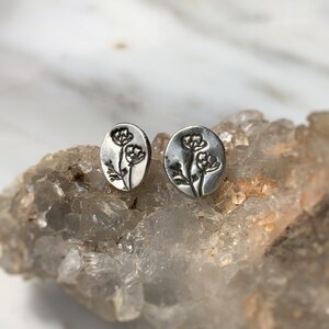 May include: A pair of silver stud earrings with a floral design. The earrings are round and have a slightly textured surface.