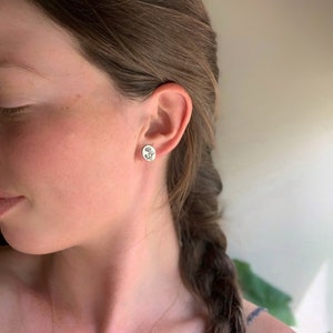 May include: A pair of silver stud earrings with a floral design. The earrings are worn in the ear of a person with long brown hair.