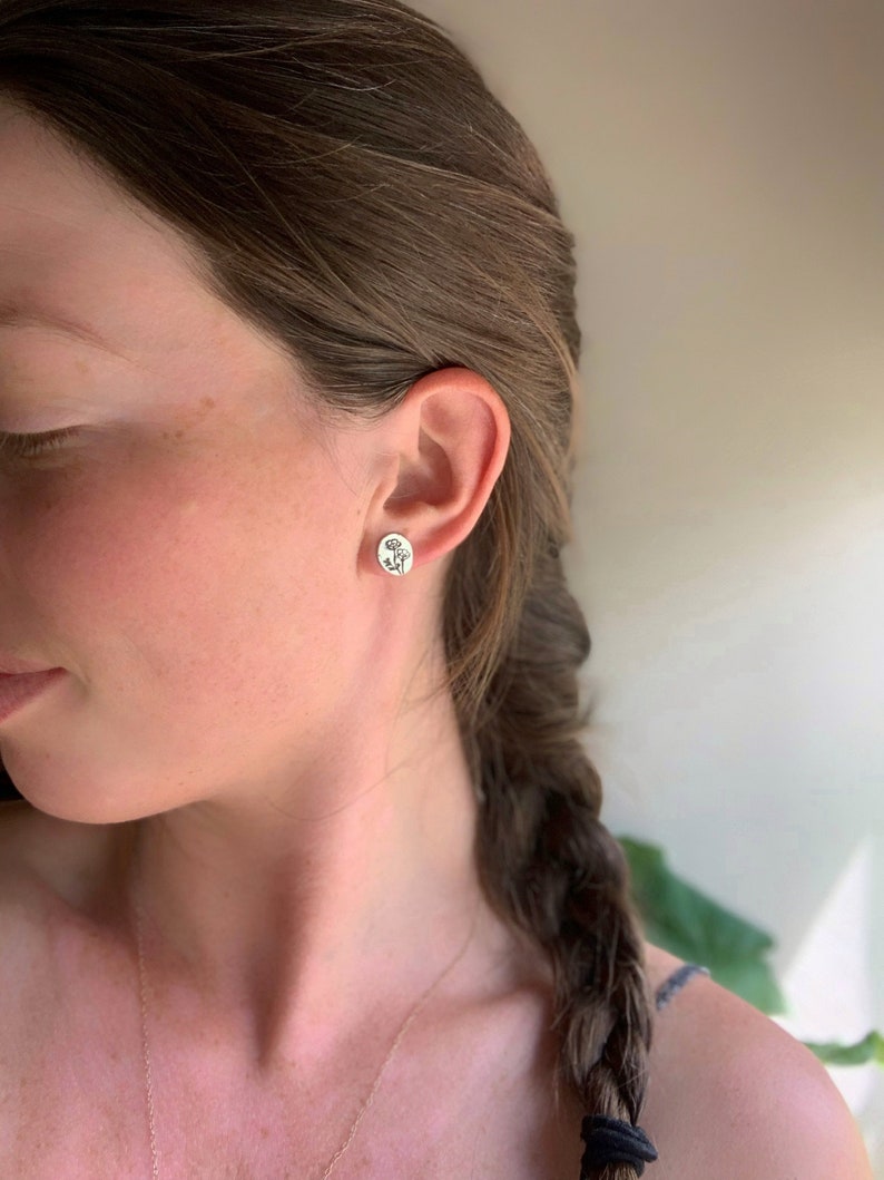 May include: A pair of silver stud earrings with a floral design. The earrings are worn in the ear of a person with long brown hair.