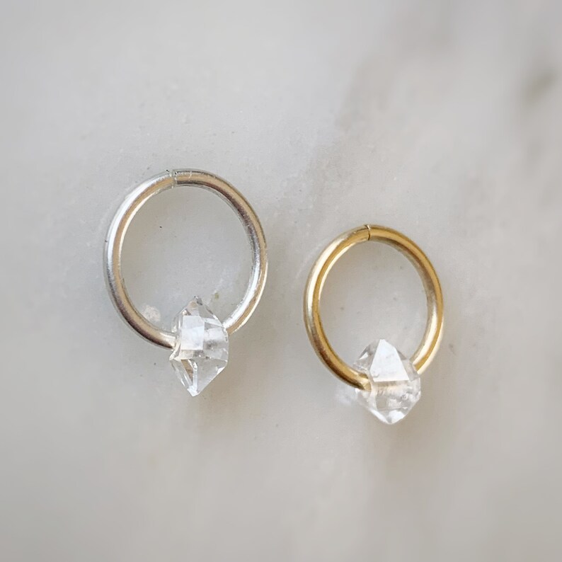 Op de afbeelding: Twee creolen, een zilveren en een gouden, elk met een helder diamantvormig kristal dat eraan hangt.