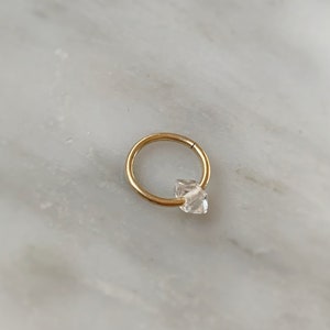 Op de afbeelding: Een gouden oorring met een heldere, gefacetteerde edelsteen. De oorring is een populaire keuze voor kraakbeenpiercings.