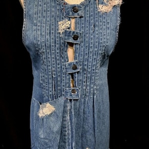 May include: A blue denim vest with a white flower embellishment, a button-up front, and a pocket on the left side. The vest has a distressed look with frayed edges and a few holes.