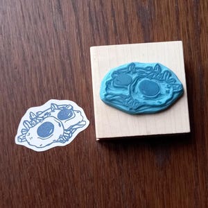 May include: A blue rubber stamp with a fried egg design, mounted on a wooden block. A matching printed image of the design is next to the stamp on a wooden surface. The stamp is a craft supply for creating egg-themed art.
