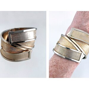 May include: A gold-toned cuff bracelet with a textured, woven appearance. The bracelet features rectangular silver-toned accents and a hinged design. The image shows the bracelet on a white background and on a wrist.