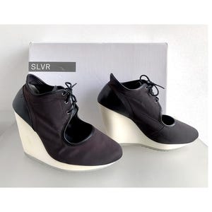 May include: A pair of black wedge heels with a cream-coloured wedge sole. The shoes feature a lace-up design and a side cutout. The heels are set against a white background.