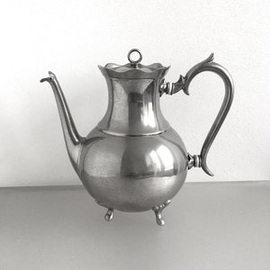 Può includere: Una teiera d'argento con corpo arrotondato e coperchio scanalato. La teiera ha un beccuccio curvo e un manico che si curva verso il corpo. La teiera poggia su tre piccoli piedini.