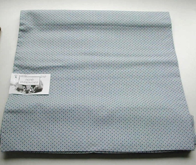 Op de afbeelding: Gevouwen lichtblauwe stof met een herhalend patroon van kleine witte en blauwe stippen. Een label met de tekst "Pugs Endeavors" is bevestigd. Geschikt voor naaien of knutselen.