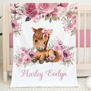 Peut inclure: Une couverture de bébé blanche avec une illustration à l'aquarelle d'un poulain brun avec un nœud rose, entouré de roses roses et de verdure. La couverture porte le nom "Marley Evelyn" en écriture rose.