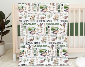 May include: A white blanket featuring a repeating pattern of mallard ducks, cattails, and the text 'Hudson James' and 'H' in various green and black fonts. The design includes illustrations of ducks in flight and swimming on water.