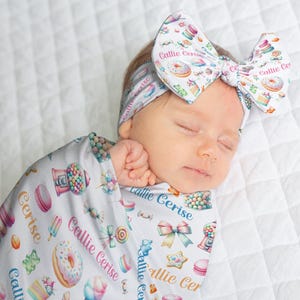 May include: A sleeping baby wrapped in a light gray swaddle and wearing a matching headband, both adorned with colorful candy and dessert illustrations. The text "Callie Cerise" is printed on the swaddle and headband.