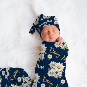 K&ouml;nnte beinhalten: Ein marineblauer Swaddle mit einem wei&szlig;en G&auml;nsebl&uuml;mchenmuster und dem Namen "Daisy Mae" in goldenen Buchstaben. Eine passende marineblaue und wei&szlig;e G&auml;nsebl&uuml;mchenm&uuml;tze mit dem Namen "Daisy Mae" in goldenen Buchstaben befindet sich auf dem Kopf des Babys.