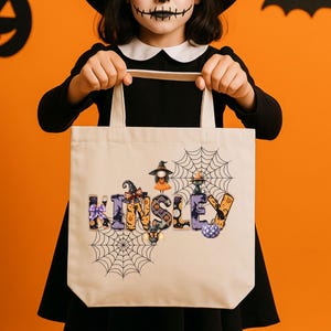 May include: A beige Halloween tote bag with the name "KINSLEY" in colorful, spooky lettering. The bag features spiderwebs and Halloween-themed illustrations. The background is orange with a pumpkin and bat cutouts.