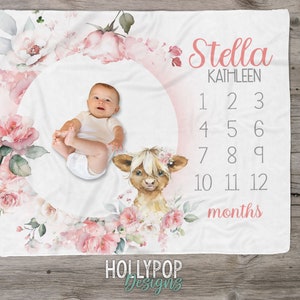 May include: A white baby blanket with a pink floral design and a circle in the center. The circle has a baby's photo, a highland cow illustration, and numbers 1-12 for monthly milestones. The text "Stella Kathleen" and "months" are also on the blanket.