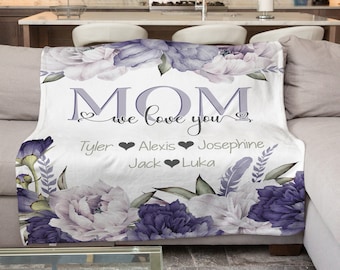 May include: A white fleece blanket with a floral design in shades of purple and white. The blanket features the text "MOM we love you" with a heart on each side of the text. Below the text are the names "Tyler, Alexis, Josephine, Jack, Luka" with a heart between each name.