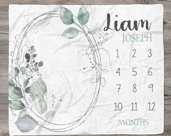 Puede incluir: Una manta de bebé blanca con un diseño de corona de acuarela gris y verde. La manta tiene una tabla de números para hacer un seguimiento del crecimiento de un bebé, con el nombre "Liam Joseph" en la parte superior. El texto "Hollypop Designs" está en la parte inferior de la manta.