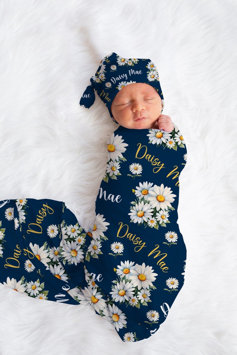 K&ouml;nnte beinhalten: Ein marineblauer Swaddle mit einem wei&szlig;en G&auml;nsebl&uuml;mchenmuster und dem Namen "Daisy Mae" in goldenen Buchstaben. Eine passende marineblaue und wei&szlig;e G&auml;nsebl&uuml;mchenm&uuml;tze mit dem Namen "Daisy Mae" in goldenen Buchstaben befindet sich auf dem Kopf des Babys.