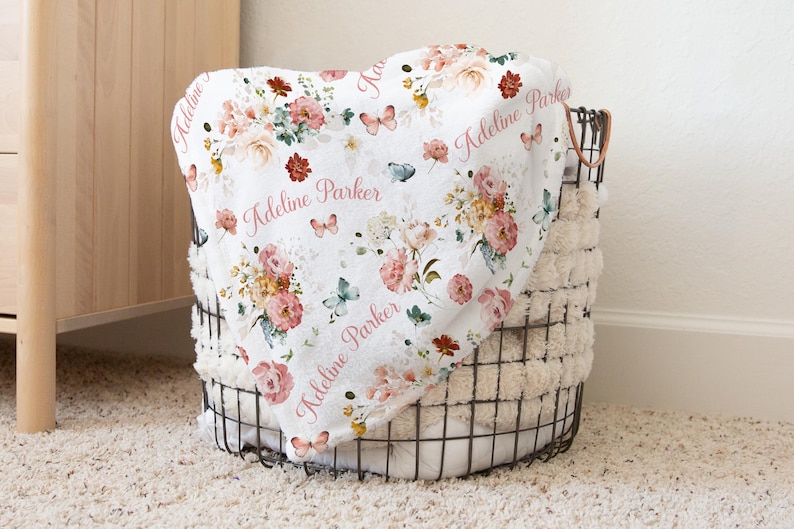 May include: A white floral baby blanket with pink roses, butterflies, and the name "Adeline Parker" printed in pink. The blanket is folded in a black wire basket with a fluffy white lining, sitting on a cream-colored carpet.
