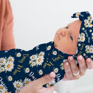 K&ouml;nnte beinhalten: Ein marineblauer Swaddle mit einem wei&szlig;en G&auml;nsebl&uuml;mchen-Druck und dem Namen "Daisy Mae", der sich &uuml;ber die gesamte Oberfl&auml;che wiederholt. Ein passendes Stirnband mit dem gleichen Druck ist um den Kopf des Babys gebunden.