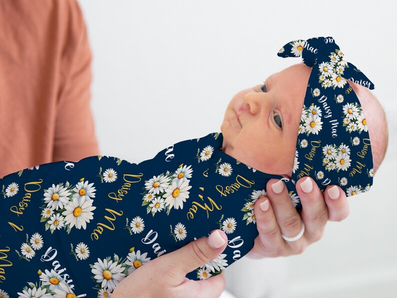 K&ouml;nnte beinhalten: Ein marineblauer Swaddle mit einem wei&szlig;en G&auml;nsebl&uuml;mchen-Druck und dem Namen "Daisy Mae", der sich &uuml;ber die gesamte Oberfl&auml;che wiederholt. Ein passendes Stirnband mit dem gleichen Druck ist um den Kopf des Babys gebunden.