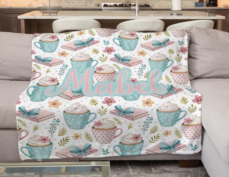 May include: A white blanket with a repeating pattern of blue polka-dotted coffee cups, pink books tied with teal bows, and floral accents. The name "Mabel" is written in pink and teal script across the blanket.