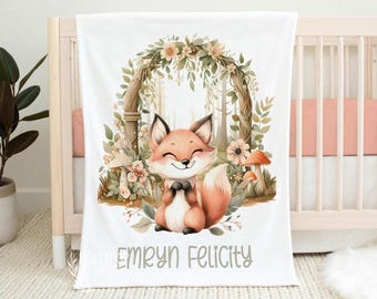 May include: A white baby blanket featuring a central illustration of a smiling fox sitting under a floral archway with mushrooms. The name Emryn Felicity is printed in gray text at the bottom of the blanket, which is draped over a wooden crib.