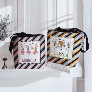 May include: Two Halloween-themed tote bags with black handles. One bag features a pink, gray, and black striped pattern with a framed image of skeleton ballerinas and the name "MICHAELA". The other bag has an orange, gray, and black striped pattern with skeleton dancers and the name "LINCOLN".