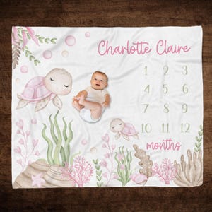 May include: A white baby blanket with a pink and green underwater theme. It features two pink sea turtles, coral, seaweed, and bubbles. The blanket has a personalized name "Charlotte Claire" and a number chart for tracking a baby's growth from 1 to 12 months.