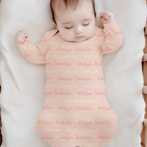 May include: A baby wearing a peach-colored sleep sack with the name "Paityn Isabella" printed repeatedly in a light pink script. The baby is lying in a wooden crib on a white blanket. The sleep sack has long sleeves and a footed design.