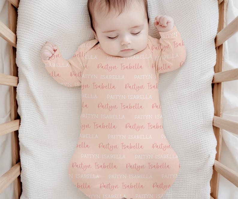May include: A baby wearing a peach-colored sleep sack with the name "Paityn Isabella" printed repeatedly in a light pink script. The baby is lying in a wooden crib on a white blanket. The sleep sack has long sleeves and a footed design.