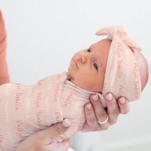 May include: A newborn baby wrapped in a light pink swaddle and matching headband. The fabric is printed with the name "Paityn Isabella" in a repeating pattern. The baby is held gently in someone's hands.