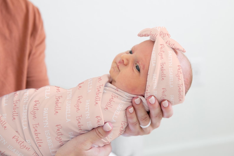 May include: A newborn baby wrapped in a light pink swaddle and matching headband. The fabric is printed with the name "Paityn Isabella" in a repeating pattern. The baby is held gently in someone's hands.