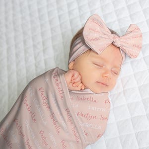 May include: A newborn baby wrapped in a pink swaddle blanket and wearing a matching headband with a bow. The blanket and headband are printed with the names "Paityn" and "Isabella". The baby is lying on a white quilted surface.
