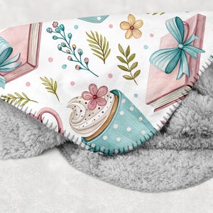 May include: A soft, gray and white blanket with a whimsical pattern. The pattern includes pink books tied with blue bows, a blue polka-dot cup with a flower, and floral accents. The blanket has a plush, fuzzy texture.
