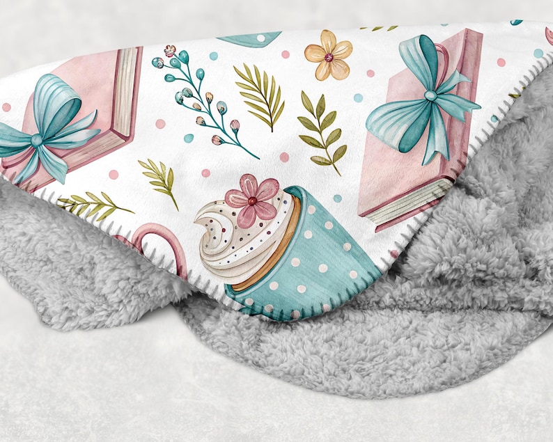 May include: A soft, gray and white blanket with a whimsical pattern. The pattern includes pink books tied with blue bows, a blue polka-dot cup with a flower, and floral accents. The blanket has a plush, fuzzy texture.