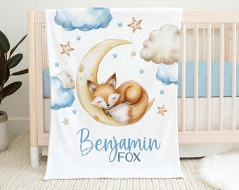 May include: A white baby blanket featuring a sleeping fox on a crescent moon, surrounded by blue clouds and stars. The name "Benjamin FOX" is written in blue script. The blanket is displayed on a crib.