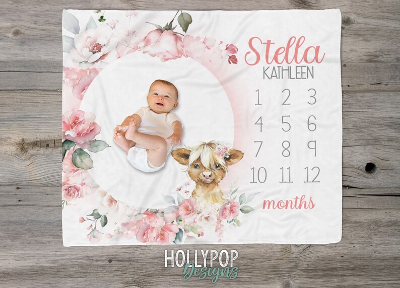 May include: A white baby blanket with a pink floral design and a circle in the center. The circle has a baby's photo, a highland cow illustration, and numbers 1-12 for monthly milestones. The text "Stella Kathleen" and "months" are also on the blanket.