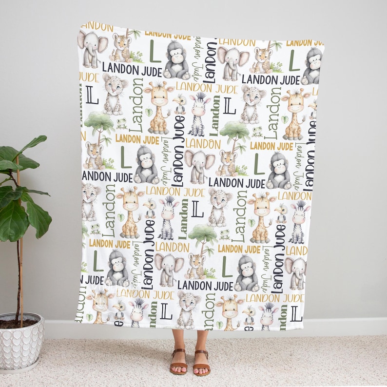 A person is standing in front of a white blanket with a repeating pattern of various animals, including elephants, giraffes, and zebras, along with the name 'Landon' written on it.