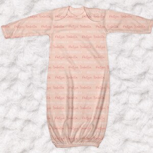May include: A light peach-colored baby gown with long sleeves. The gown is covered in the repeating text "Paityn Isabella" in a soft pink hue. The gown is made of a soft material and is designed for newborns.