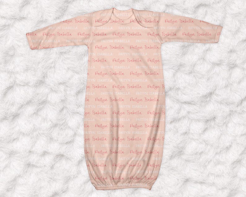 May include: A light peach-colored baby gown with long sleeves. The gown is covered in the repeating text "Paityn Isabella" in a soft pink hue. The gown is made of a soft material and is designed for newborns.