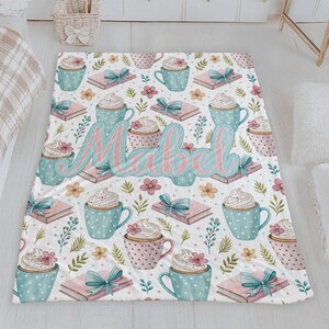 May include: A white blanket with a repeating pattern of blue and pink polka-dot mugs topped with whipped cream, pink books with blue bows, and flowers. The name "Mabel" is written in large, cursive letters across the blanket.