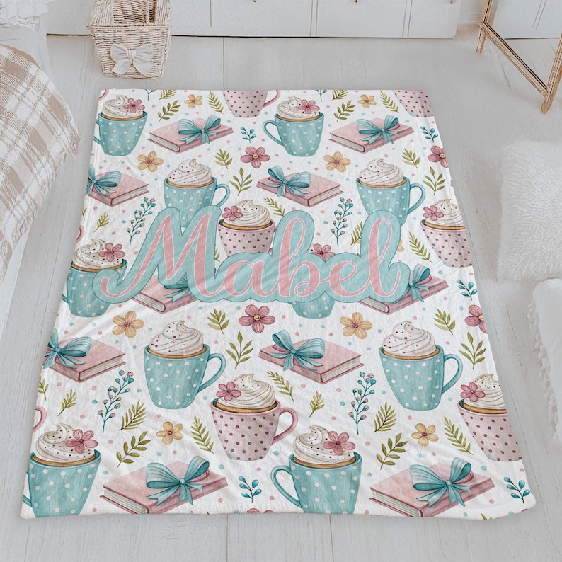May include: A white blanket with a repeating pattern of blue and pink polka-dot mugs topped with whipped cream, pink books with blue bows, and flowers. The name "Mabel" is written in large, cursive letters across the blanket.