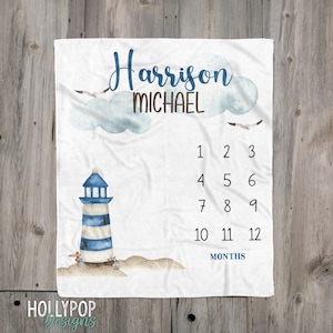 May include: A white baby blanket with a blue and white lighthouse illustration and a number chart for tracking a baby's growth. The blanket has the name "Harrison Michael" written on it.