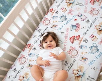 May include: A white crib sheet featuring a repeating pattern of illustrations including a girl with blonde hair, a blue teapot, a pocket watch, a cupcake, a cat, and the name Alice Kathleen printed in a cursive script font.