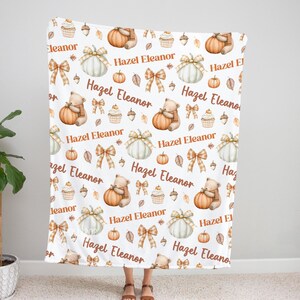 May include: A white blanket with a repeating pattern of pumpkins, bows, teddy bears, cupcakes, and acorns. The blanket has the text "Hazel Eleanor" in a brown script. The blanket is held up by a person.